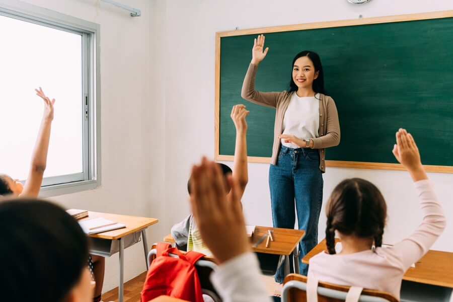 Asian school teacher with students raising hands.